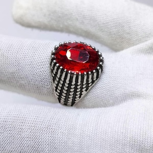 May include: A silver-toned ring featuring a large, oval-cut, vibrant red gemstone set in a prominent, textured metal band. The band displays a series of vertical, grooved lines that extend from the base of the setting down the sides of the ring.
