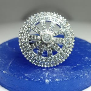 May include: A silver ring with a circular design, set with small, clear gemstones. The centre of the ring features a larger gemstone. The ring is displayed on a blue surface.