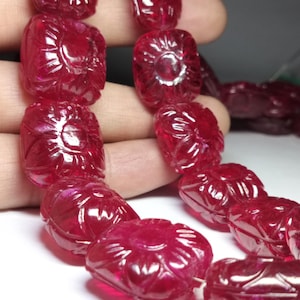 Könnte beinhalten: Eine Kette aus geschnitzten Rubinperlen. Jede Perle hat eine tiefrote Farbe und weist ein Blumendesign auf. Die Perlen sind aufgefädelt und bilden eine Halskette oder ein Armband. Die aufwendigen Schnitzereien und die satte Farbe machen diese Perlen zu einem auffälligen Schmuckstück.