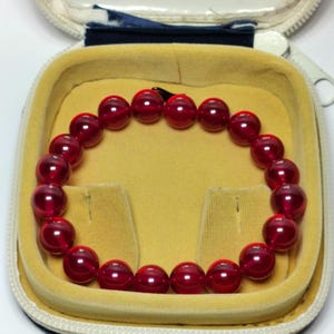 May include: A circular bracelet composed of smooth, translucent deep red spherical beads. The beads are uniform in size and strung together to form a continuous loop, resting inside a small, open, beige-lined protective storage case.