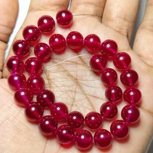 May include: A strand of round, translucent, deep red beads with a distinct cat's eye effect, featuring a bright horizontal light reflection across the center of each bead. The beads are strung on a thin, flexible wire and arranged in a circular shape.
