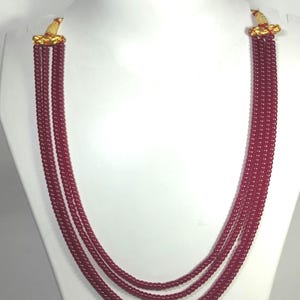 May include: A multi-strand necklace with small, round, burgundy beads. The necklace has gold-colored decorative clasps at the top. The necklace is displayed on a white mannequin bust. The necklace is a jewelry item.