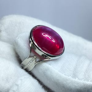 Puede incluir: Un anillo de plata con una gran piedra preciosa cabujón lisa de forma ovalada en un color rojo magenta intenso y vibrante. La piedra está engastada en un bisel de plata pulida y la banda del anillo tiene un diseño dividido.