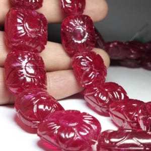 Könnte beinhalten: Eine Kette aus geschnitzten Rubinperlen. Jede Perle hat eine tiefrote Farbe und weist ein Blumendesign auf. Die Perlen sind aufgereiht und zeigen ihre komplizierten Details und polierten Oberflächen. Die Edelsteine werden in einer Hand gehalten.