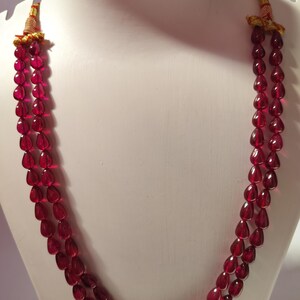 May include: A two-strand necklace featuring teardrop-shaped, translucent red beads. The necklace is displayed on a white mannequin bust. The necklace's ends are adorned with gold-colored tassels.