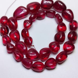 May include: A strand of polished, translucent deep red gemstone beads with an irregular, rounded pebble shape. Each bead features a smooth, glossy surface that reflects light, arranged in a circular pattern on a thin, light-coloured string.