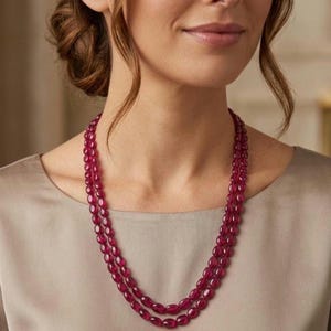 May include: A double-strand necklace featuring smooth, oval-shaped beads in a deep, saturated ruby red colour. The beads are uniform in size and shape, strung together to create two distinct layers that drape gracefully around the neck area.