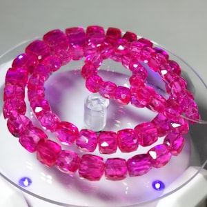 May include: A strand of faceted, hot pink beads. The beads are square-shaped with multiple facets, creating a sparkling effect. The beads are strung together, forming a circular shape. The background is a light, reflective surface.