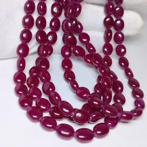 May include: A strand of smooth, polished, deep magenta-coloured oval beads. The beads are uniform in shape and size, strung together to form a continuous loop. The surface of each bead reflects light, highlighting the rich, saturated berry tone.