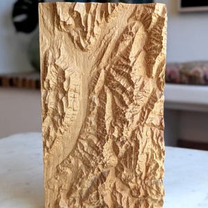 Puede incluir: Un bloque de madera rectangular con un detallado relieve de un paisaje montañoso. La madera es de color marrón claro natural, y el relieve representa valles y picos. La pieza se exhibe sobre una superficie blanca.