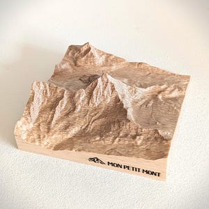 Puede incluir: Escultura en relieve de madera de color marrón claro que representa una cordillera. La escultura presenta tallas detalladas de picos y valles. El texto "MON PETIT MONT" está impreso en la base.