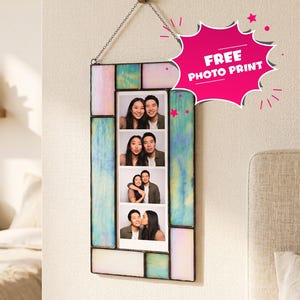 A rectangular stained glass photobooth frame with iridescent pastel panels in shades of blue, pink, and white. The frame hangs from a metal chain on a wall hook and contains a vertical photo strip showing a couple in four poses. A pink starburst graphic with the text FREE PHOTO PRINT is attached.