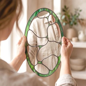 May include: An oval-shaped mirror featuring a decorative stained glass border in shades of green. The frame is constructed with dark metal soldering that creates an organic, irregular pattern of glass segments surrounding the central reflective surface.
