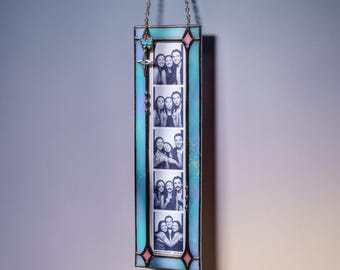May include: A rectangular stained glass photo frame with a light blue and teal border, featuring pink diamond accents at the corners. A vertical strip of four black and white photo booth pictures is displayed inside, with a small blue butterfly charm.