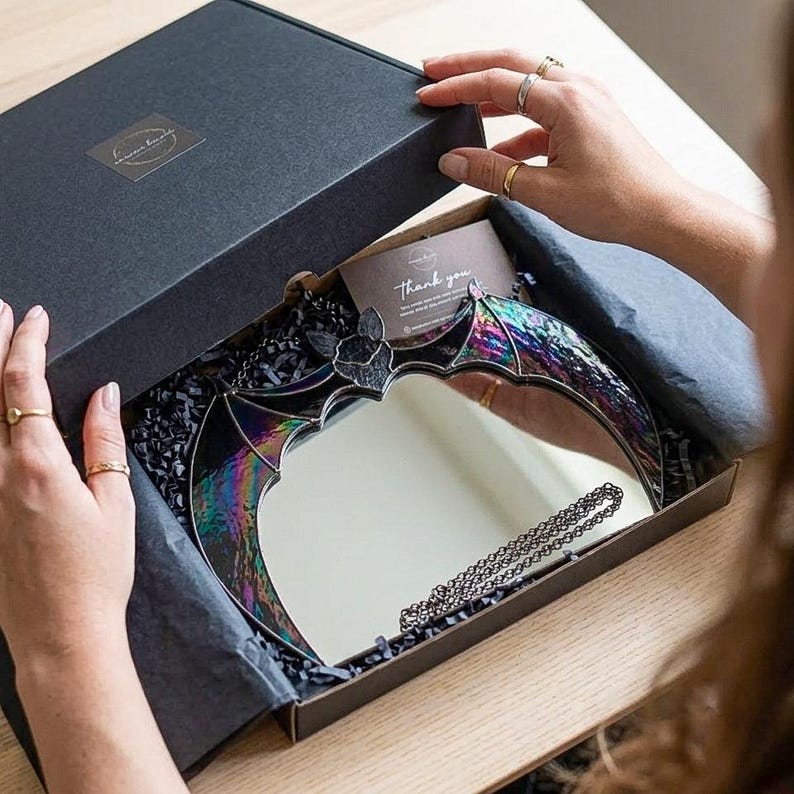 May include: A bat-shaped mirror with iridescent stained glass wings in a black gift box. The mirror features a central reflective surface framed by dark, shimmering glass panels. A small thank you card is tucked inside the box with black tissue paper.