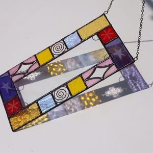 May include: A rectangular stained glass suncatcher featuring a border of multicolored glass panes including yellow, pink, blue, purple, and red segments. The design incorporates etched star, spiral, and floral patterns, all held together by dark metal solder lines.