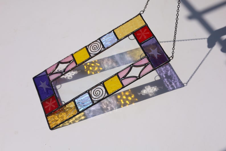 May include: A rectangular stained glass suncatcher featuring a border of multicolored glass panes including yellow, pink, blue, purple, and red segments. The design incorporates etched star, spiral, and floral patterns, all held together by dark metal solder lines.