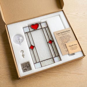 May include: A rectangular stained glass panel featuring a red heart at the top and red diamond accents, framed in dark metal. The set includes a suction cup, a metal hook, a silver chain, and care instruction cards in a cardboard box.