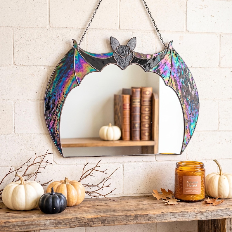 A wall-mounted stained glass mirror shaped like a bat with iridescent, multi-colored stained glass wings and a central bat head. The mirror is suspended by a black metal chain and features a reflective surface that shows a reflection of books.