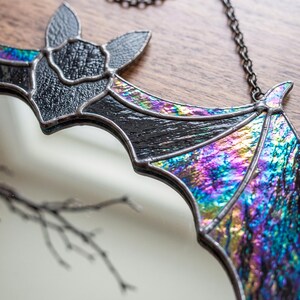 May include: A stained glass bat suncatcher featuring textured black glass for the body and iridescent, multi-colored glass with purple, blue, and green hues for the wings. The piece is held together by dark metal solder lines and hangs from a chain.
