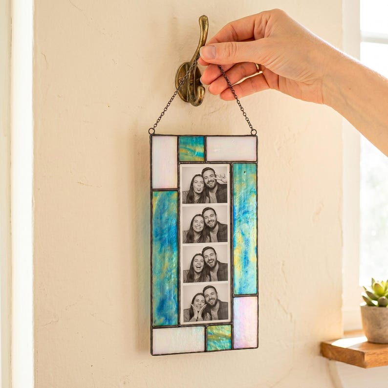 May include: A rectangular stained glass photo frame featuring iridescent white and blue-green glass panels. The frame holds a vertical strip of four black and white photos of a couple and is suspended by a metal chain from a wall hook.