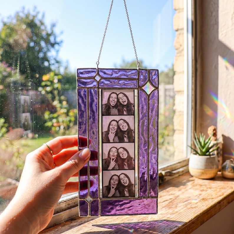 May include: A rectangular stained glass photo frame with a vibrant purple, textured border and a metal chain for hanging. The frame holds a vertical strip of four black and white photo booth pictures showing two people making various facial expressions.