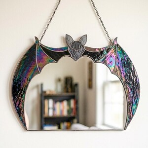 May include: A bat-shaped wall mirror featuring iridescent, multi-colored stained glass wings and a dark, textured glass head. The mirror is framed in dark metal solder and hangs from a thin metal chain, creating a crescent-like silhouette.