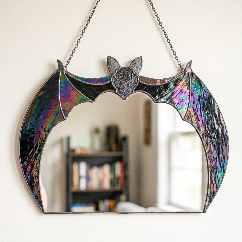 May include: A bat-shaped wall mirror featuring iridescent, multi-colored stained glass wings and a dark, textured glass head. The mirror is framed in dark metal solder and hangs from a thin metal chain, creating a crescent-like silhouette.