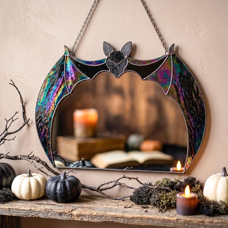 A wall-hanging stained glass mirror shaped like a flying bat with iridescent, multi-colored stained glass wings and a black glass body. The mirror is suspended by a thin metal chain and features a reflective surface that shows a blurred interior scene.