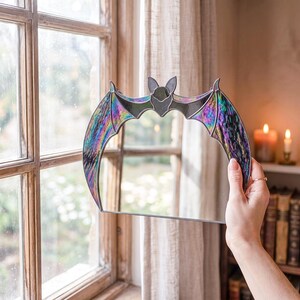 A stained glass bat mirror featuring iridescent, multi-colored stained glass wings with a dark, reflective center. The glass is framed in thin metal, creating a crescent-like silhouette with pointed ears at the top center of the frame.