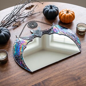 A bat-shaped wall mirror featuring iridescent, multi-colored stained glass wings and a central reflective surface. The mirror is framed in dark metal solder and includes a matching metal chain for hanging, showcasing a reflective finish.