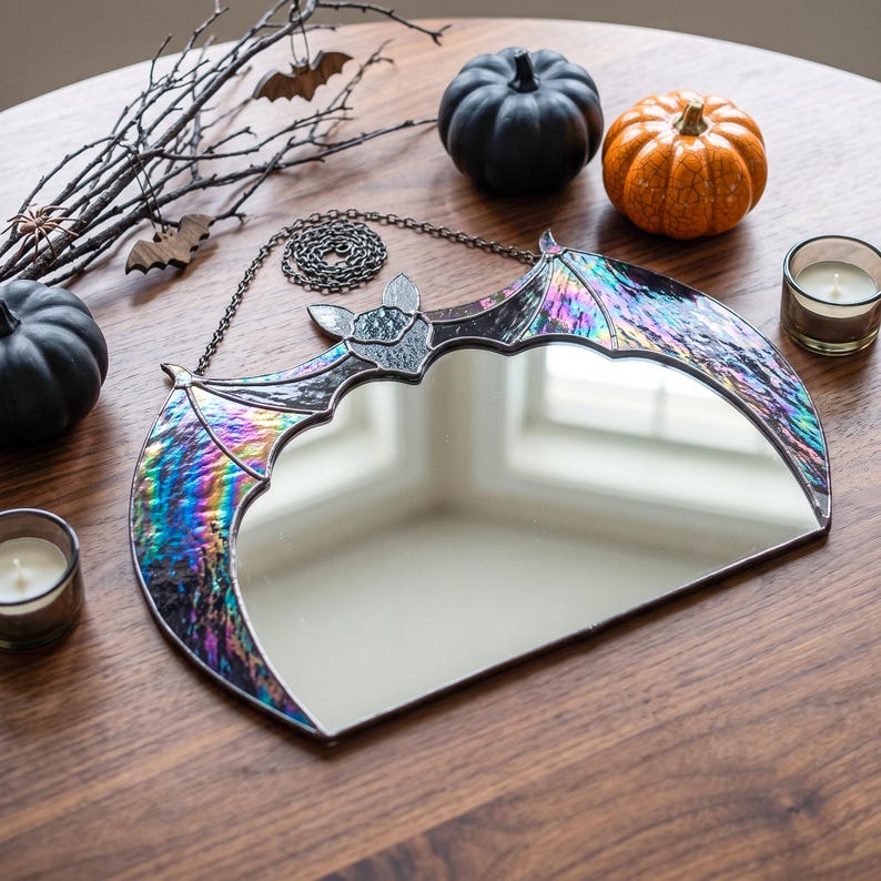 A bat-shaped wall mirror featuring iridescent, multi-colored stained glass wings and a central reflective surface. The mirror is framed in dark metal solder and includes a matching metal chain for hanging, showcasing a reflective finish.