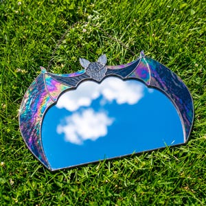 May include: A bat-shaped wall mirror featuring iridescent, multi-colored stained glass wings that shimmer with purple, blue, and green hues. The central mirror reflects the sky and clouds, and the piece includes a thin metal hanging chain.