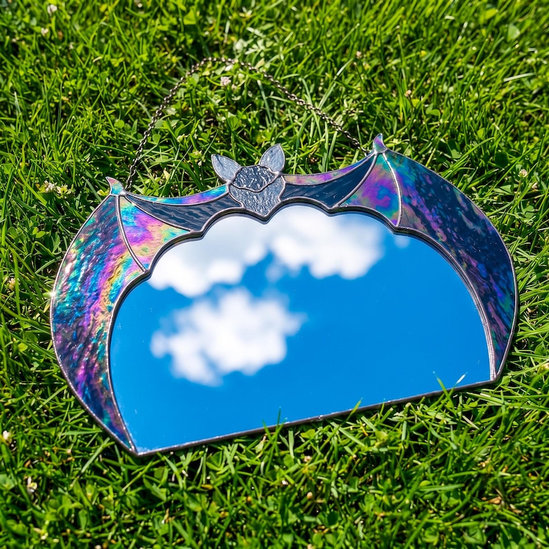 May include: A bat-shaped wall mirror featuring iridescent, multi-colored stained glass wings that shimmer with purple, blue, and green hues. The central mirror reflects the sky and clouds, and the piece includes a thin metal hanging chain.