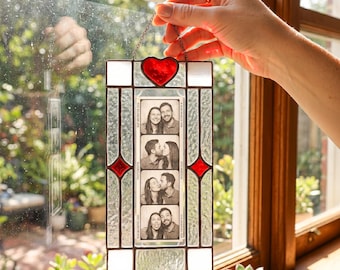 May include: A rectangular stained glass frame with a red heart at the top and red diamond accents on the sides. The frame holds a vertical black and white photo strip featuring four images of a couple. The glass has a textured finish.