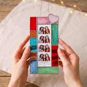 May include: A rectangular stained glass photo frame featuring a vertical strip of four photos of three people. The frame has multicolored glass panels in red, pink, blue, and green, with a small heart-shaped glass piece at the top. The photo strip includes the text "SUMMER MEMORIES 2024".