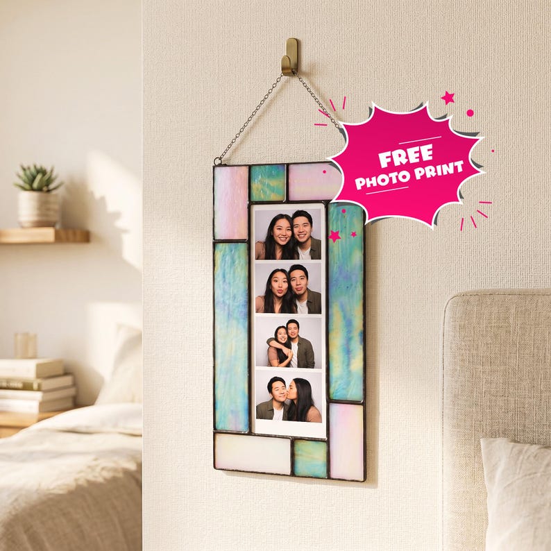 A rectangular stained glass photobooth frame with iridescent pastel panels in shades of blue, pink, and white. The frame hangs from a metal chain on a wall hook and contains a vertical photo strip showing a couple in four poses. A pink starburst graphic with the text FREE PHOTO PRINT is attached.