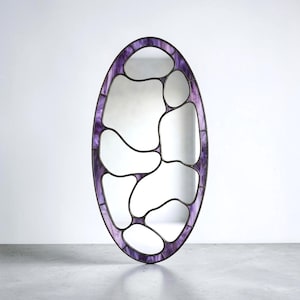 May include: An oval-shaped mirror featuring a frame constructed from purple marbled glass segments joined by dark metal lines. The interior of the mirror is divided into multiple irregular, organic-shaped sections by the dark metal framework.