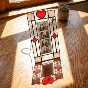 May include: A rectangular stained glass photo frame featuring a central vertical strip of four black and white photo booth images of a couple. The frame has a red glass heart at the top, red diamond accents, and iridescent textured glass panels.
