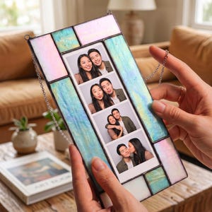A rectangular stained glass photo frame featuring iridescent pastel blue, pink, and white glass panels. The frame holds a vertical strip of four black and white photo booth pictures showing a couple in various poses, suspended by a metal chain.