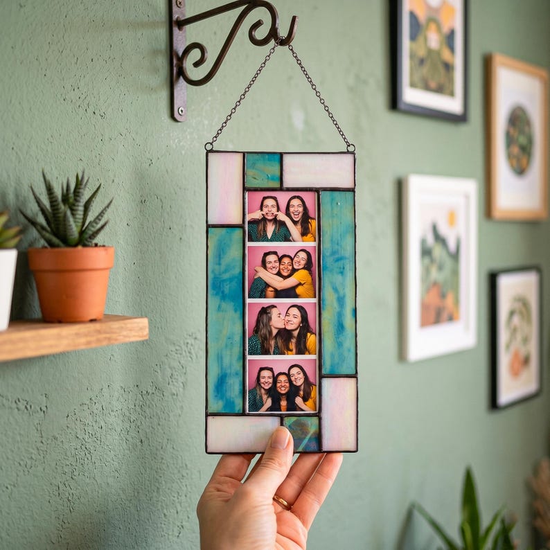A rectangular stained glass photobooth picture frame constructed from stained glass pieces in shades of teal and iridescent white, held together by black patina solder lines. A hand holds the frame displaying a custom photo strip of friends, hanging on a textured green wall.