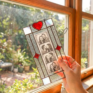 May include: A rectangular stained glass photo frame with a red heart at the top and red diamond accents. The frame holds a vertical black and white photo strip featuring four different poses of a couple. The glass has a textured, iridescent finish.