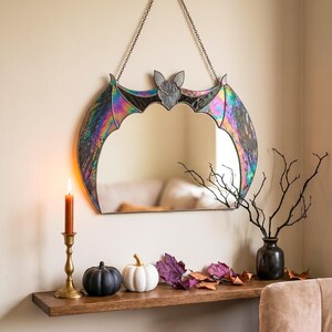 May include: A wall-mounted mirror shaped like a bat with iridescent stained glass wings and a central bat head. The mirror is suspended by a dark metal chain and features a reflective surface framed by dark metal edges and colorful glass panels.