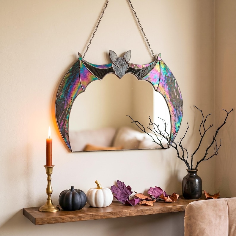 May include: A wall-mounted mirror shaped like a bat with iridescent stained glass wings and a central bat head. The mirror is suspended by a dark metal chain and features a reflective surface framed by dark metal edges and colorful glass panels.