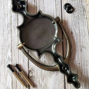 Puede incluir: Un llamador de puerta negro y plateado con un dise&ntilde;o decorativo. Tiene una placa redonda con un dise&ntilde;o en relieve y un asa curva. El llamador viene con dos tornillos y dos arandelas de goma negras.