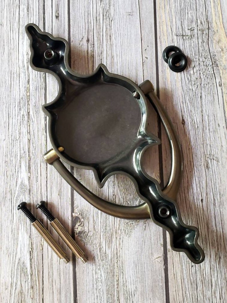 Puede incluir: Un llamador de puerta negro y plateado con un dise&ntilde;o decorativo. Tiene una placa redonda con un dise&ntilde;o en relieve y un asa curva. El llamador viene con dos tornillos y dos arandelas de goma negras.