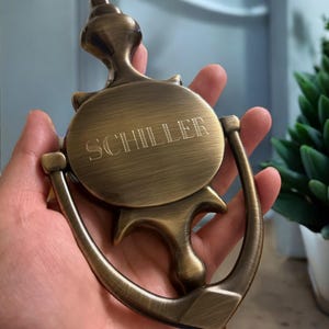 Puede incluir: Un llamador de puerta de lat&oacute;n con una placa redonda grabada con el nombre "SCHILLER".