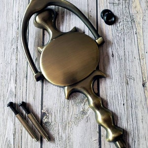 Puede incluir: Un llamador de puerta de lat&oacute;n con un dise&ntilde;o ornamentado. El llamador tiene una placa circular con un borde elevado y un mango largo y curvo. El llamador est&aacute; montado en una superficie de madera.