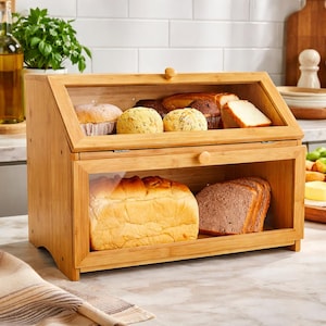 May include: A two-tiered bamboo bread box with clear glass doors. The top section holds muffins and rolls, while the bottom section contains a loaf of white bread and sliced brown bread. The box has a wooden knob for opening.