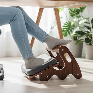 May include: A wooden footrest with a gray cushion and a curved, ergonomic design. The footrest is under a desk, with a person's feet resting on it. The frame is dark brown, and the cushion is gray.